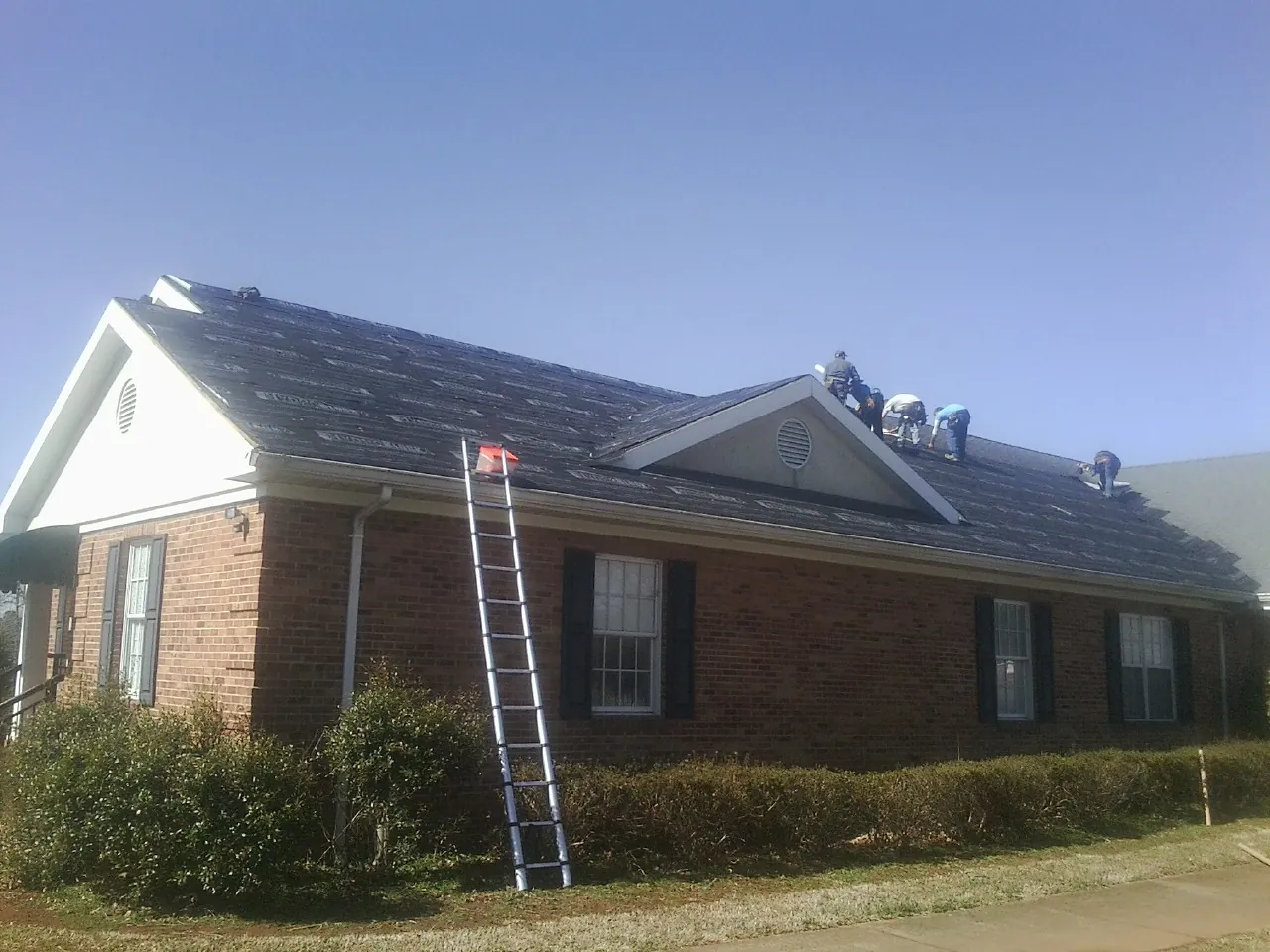 Crew tearing shingles off a brick ranch roof with a ladder set — Metal Roof Repair work by our crew in Jessup